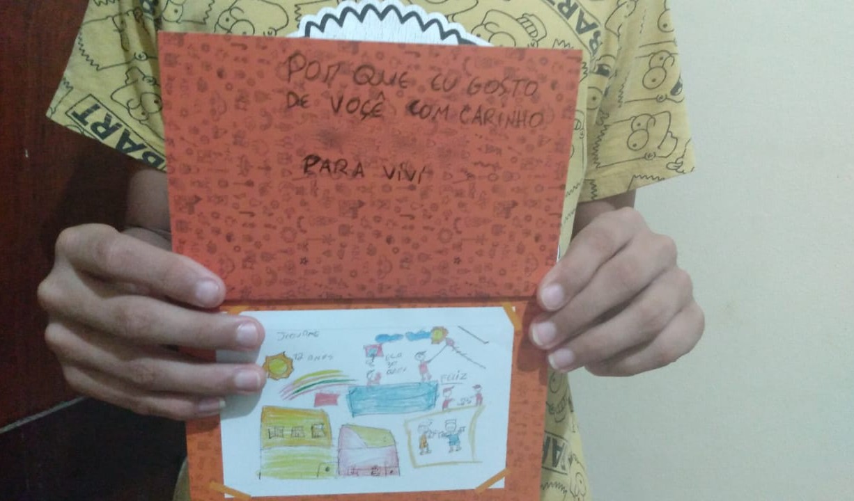 a foto mostra as mãos de um menino, que não mostra o rosto segurando um desenho que tem duas casas, uma amarela e outra rosa, o sol, um arco-íris e crianças brincando